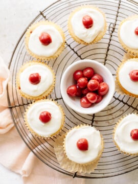 Vanilla cupcakes with white icing and cherries on top, arranged on a cooling rack with a bowl of cherries in the center.