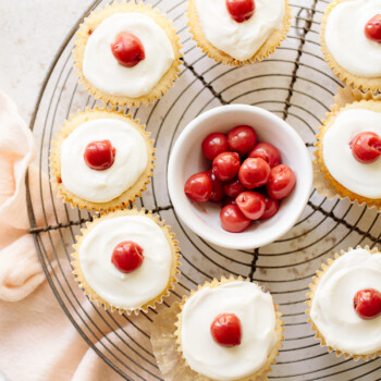 Vanilla cupcakes with white icing and cherries on top, arranged on a cooling rack with a bowl of cherries in the center.