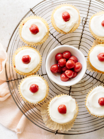 Vanilla cupcakes with white icing and cherries on top, arranged on a cooling rack with a bowl of cherries in the center.