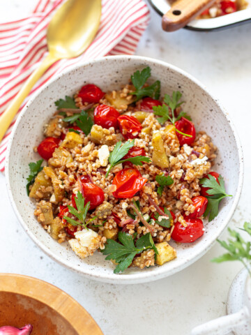 A vibrant farro bowl with cherry tomatoes, parsley, eggplant pieces, and feta cheese sits on a white surface, accompanied by a gold spoon and a red striped napkin.