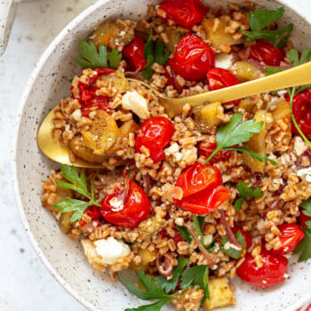 A bowl of cooked farro grains with roasted cherry tomatoes, parsley, feta cheese, and chunks of eggplant is served with a gold spoon resting inside.