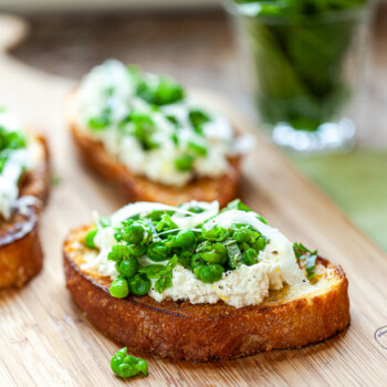 Three slices of toasted bread topped with ricotta cheese, green peas, herbs, and thinly sliced onions create a vibrant pea bruschetta on a wooden board. A glass with fresh green herbs is blurred in the background.