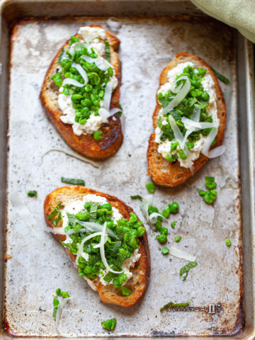 Three slices of toasted bread topped with ricotta cheese, chopped green herbs, peas, and shredded cheese create a vibrant pea bruschetta, all beautifully arranged on a metal baking tray.
