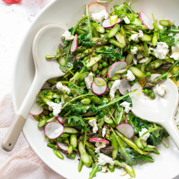 A serving bowl with chopped asparagus, sliced radishes and goat cheese with with two salad servers.