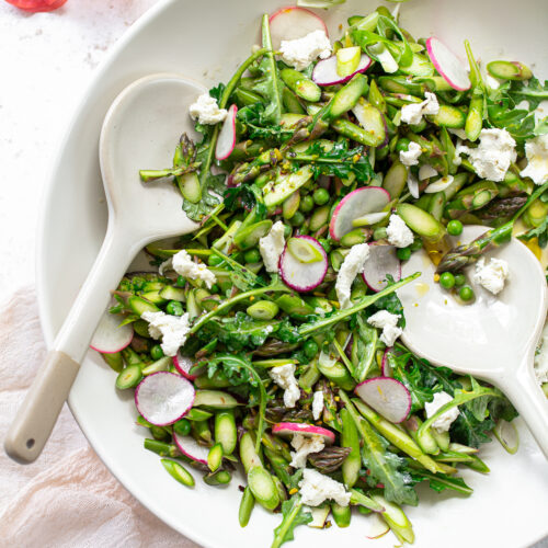 A serving bowl with chopped asparagus, sliced radishes and goat cheese with with two salad servers.