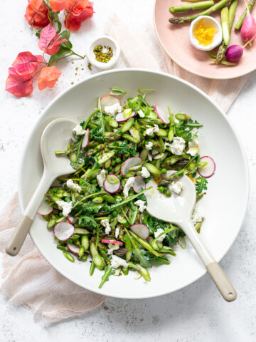 A fresh asparagus salad with radishes, leafy greens, and crumbled cheese sits in a white bowl with serving utensils. Nearby are flowers, a small bowl of seasoning, and a plate featuring asparagus and a soft-boiled egg.