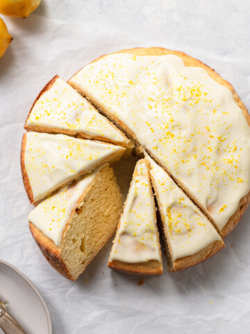 A round lemon ricotta cake with white icing and yellow zest on top, sliced into six pieces on parchment paper, with lemons and a plate visible nearby.