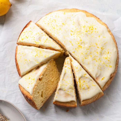A round lemon ricotta cake with white icing and yellow zest on top, sliced into six pieces on parchment paper, with lemons and a plate visible nearby.