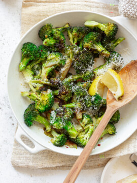 A white pan full of sauteed broccoli, with grated cheese on top and a wooden spoon and lemon wedges on the side.