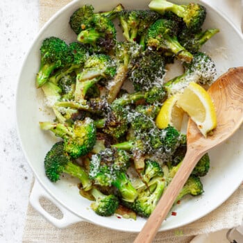 A white pan full of sauteed broccoli, with grated cheese on top and a wooden spoon and lemon wedges on the side.
