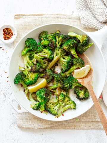A skillet with sauteed broccoli florets, lemon wedges, red chili flakes and a wooden spoon.
