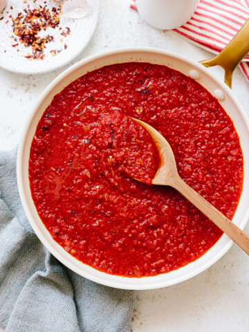 Thick tomato sauce in a skillet, with a wood spoon and bowl of chili flakes on the side.
