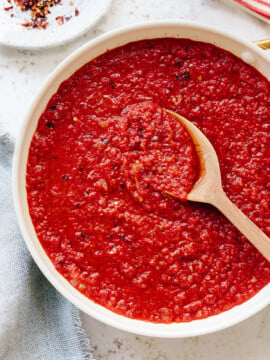 A bowl of chunky red arrabbiata sauce with visible spices, being stirred by a wooden spoon on a light-colored surface.