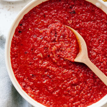 A bowl of chunky red arrabbiata sauce with visible spices, being stirred by a wooden spoon on a light-colored surface.