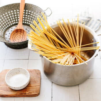 Dried spaghetti pasta in a pot with a colander and wooden spoon on a kitchen counter.