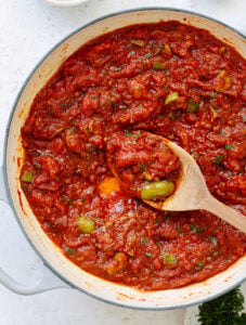A white pot filled with chunky puttanesca sauce brimming with green olives and capers, as a wooden spoon lifts some of the savory mixture.