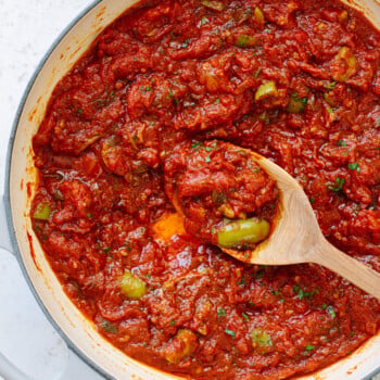 A white pot filled with chunky puttanesca sauce brimming with green olives and capers, as a wooden spoon lifts some of the savory mixture.
