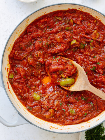 A white pot filled with chunky puttanesca sauce brimming with green olives and capers, as a wooden spoon lifts some of the savory mixture.