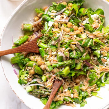 A bowl of orzo pasta salad with chopped asparagus, chickpeas, spinach, red onion, feta cheese, and herbs, tossed with a light dressing. Two wooden salad servers rest in the bowl. Pine nuts are visible in a small dish nearby.
