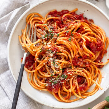 A plate of bucatini all'amatriciana topped with tomato sauce, grated cheese, and fresh herbs, with a fork resting on the side of the plate.