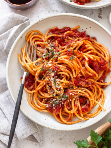 A plate of bucatini all'amatriciana topped with tomato sauce, grated cheese, and fresh herbs, with a fork resting on the side of the plate.