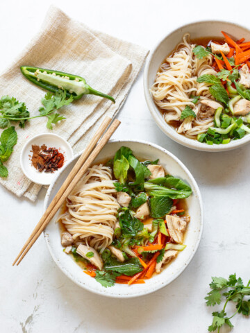 Two bowls of chicken pho soup on a surface, with chopsticks and spices on a linen napkin.