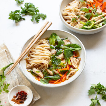 Two bowls of noodle soup with chicken, herb sprigs and chopsticks.
