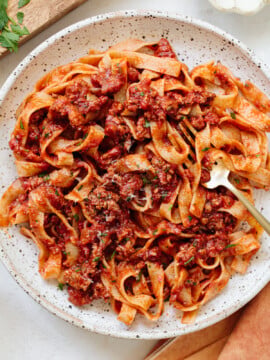 A plate of tagliatelle pasta with a hearty meat sauce, garnished with herbs. A gold fork rests on the edge of the rustic, speckled plate. The background shows a wooden board and some fresh greens.