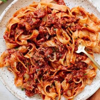 A plate of tagliatelle pasta with a hearty meat sauce, garnished with herbs. A gold fork rests on the edge of the rustic, speckled plate. The background shows a wooden board and some fresh greens.