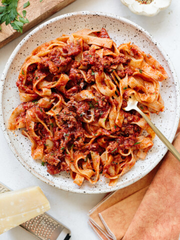 A plate of tagliatelle pasta topped with a rich, chunky tomato and meat sauce is perfect for dinner recipes. Served with a gold fork, grated cheese, a cheese block, an orange napkin, and parsley on a light tabletop.