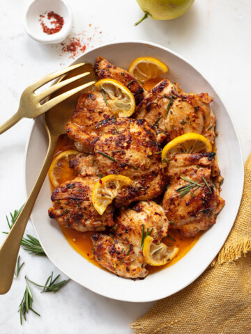 Lemon chicken thighs roasted and garnished with lemon slices and rosemary on a white oval plate, served with a gold fork and spoon, plus a yellow napkin on the side.