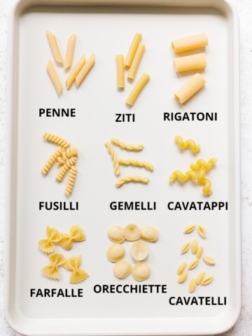 Different types of labeled pasta shapes on a white baking sheet.