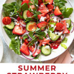 A bowl of strawberry salad with feta cheese, spinach, cherry tomatoes, cucumber slices, red onion, and herbs. The text reads Summer Strawberry Salad with Feta Cheese. Crumbled feta is in a small dish nearby.