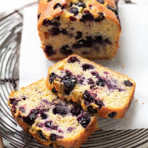 Two slices of blueberry loaf cake rest in front of a partially sliced loaf on a wire rack, showing a moist, golden interior with visible whole blueberries throughout. The bread sits on white parchment paper.