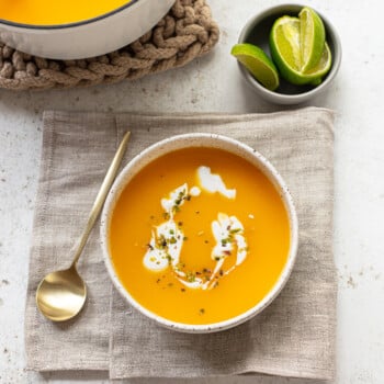 A bowl of soup on a napkin, with a spoon, dish of sliced limes and soup pot in the background.