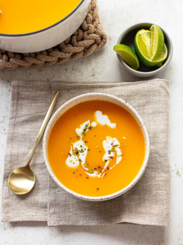 A bowl of butternut squash soup garnished with cream and herbs sits on a beige napkin with a gold spoon beside it. A small bowl of lime wedges is nearby, and part of a larger soup pot is visible at the top.