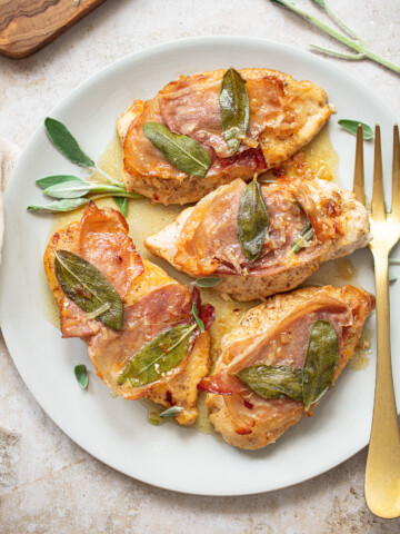 Cooked chicken breasts topped with prosciutto and sage leaves on a platter.