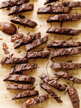 Rows of chocolate biscotti drizzled with melted chocolate are arranged on parchment paper, with some crumbs and smears of chocolate around the decadent biscotti cookies.