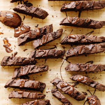 Rows of chocolate biscotti drizzled with melted chocolate are arranged on parchment paper, with some crumbs and smears of chocolate around the decadent biscotti cookies.