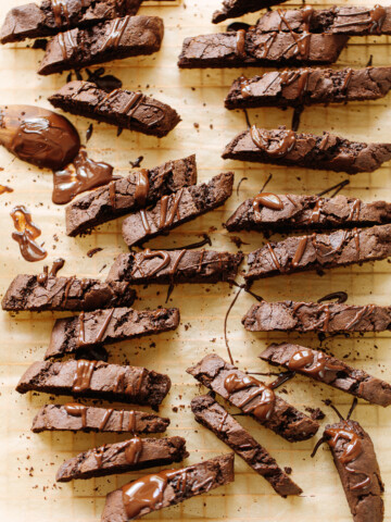 Rows of chocolate biscotti drizzled with melted chocolate are arranged on parchment paper, with some crumbs and smears of chocolate around the decadent biscotti cookies.