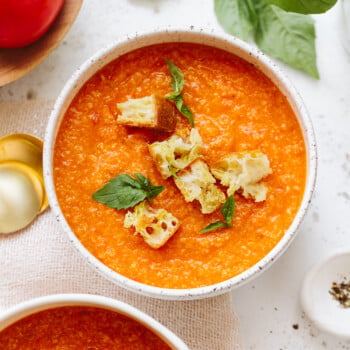 Two bowls of pappa al pomodoro tomato soup topped with croutons and fresh basil leaves, with a tomato, basil leaves, and gold spoons nearby on a white surface.