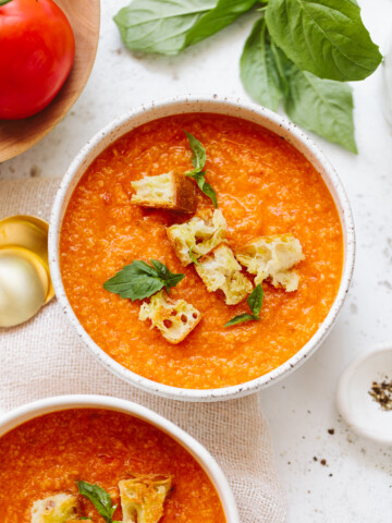 Two bowls of pappa al pomodoro tomato soup topped with croutons and fresh basil leaves, with a tomato, basil leaves, and gold spoons nearby on a white surface.
