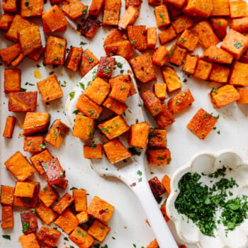 Roasted sweet potato cubes with chopped herbs are spread on a white surface, a white spoon in the center, and a small white dish of fresh chopped herbs nearby.