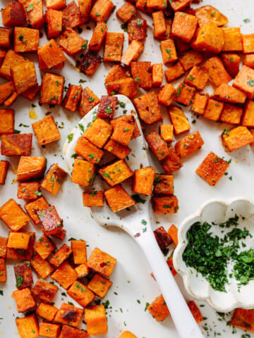 Roasted sweet potato cubes with chopped herbs are spread on a white surface, a white spoon in the center, and a small white dish of fresh chopped herbs nearby.