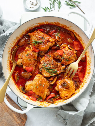 A white pot filled with chicken cacciatore—braised chicken thighs in a red tomato sauce with mushrooms, bell peppers, and fresh rosemary. Two forks rest in the pot, styled with napkins and herbs around the dish.