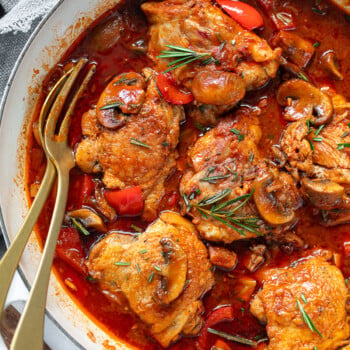 Cooked chicken thighs and tomato sauce in a braising pan, with mushrooms, rosemary sprigs and serving fork.