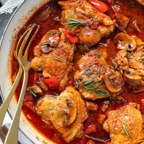 Cooked chicken thighs and tomato sauce in a braising pan, with mushrooms, rosemary sprigs and serving fork.