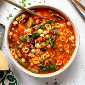 A bowl of minestrone soup with pasta shells, beans, carrots, green beans, and leafy greens in a tomato broth is served with a spoon. Chopped herbs, utensils, and parmesan cheese are nearby on a white surface.