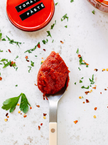 A spoonful of tomato paste with a mason jar lid on a surface sprinkled with parsley and crushed red chili.