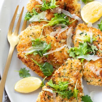 Browned chicken cutlets on a platter, with herb sprigs, shaved Parmesan and lemon wedges.